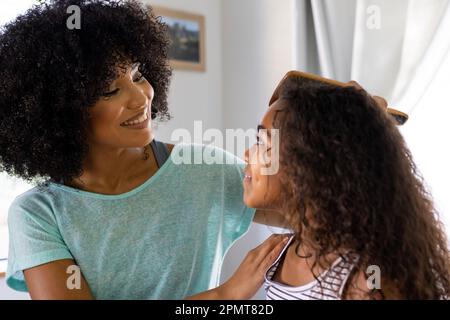 Felice madre biraciale spazzolando i capelli di sua figlia e sorridendogli in camera da letto Foto Stock