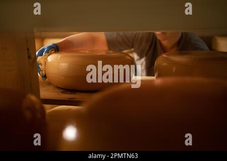 Lavoratore maschio che riveste le ruote di formaggio con cera nel magazzino di fabbrica. Teste di formaggio fresco su rack Foto Stock