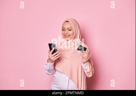 Affascinante donna musulmana in hijab rosa, con carta di credito e smartphone, effettua pagamenti via internet mobile banking Foto Stock