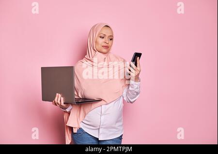 Perplesso musulmano donna incinta in hijab, tenendo il portatile e il telefono cellulare, sfondo isolato. Gravidanza. Carriera. Lavoro Foto Stock