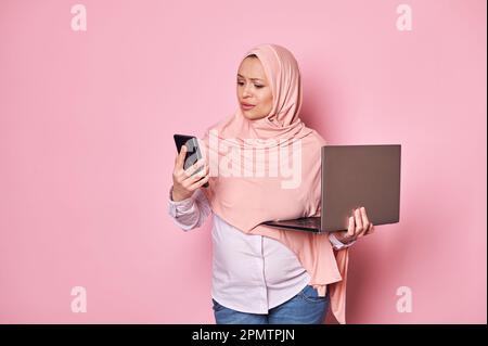 Perplesso musulmano arabo donna incinta in hijab, guardando il suo smartphone, in posa con il portatile su sfondo di colore rosa Foto Stock