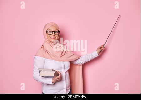 Insegnante di donna musulmana incinta in hijab rosa e occhiali, sorridendo alla macchina fotografica, indicando lo spazio copia, spiegando la lezione Foto Stock