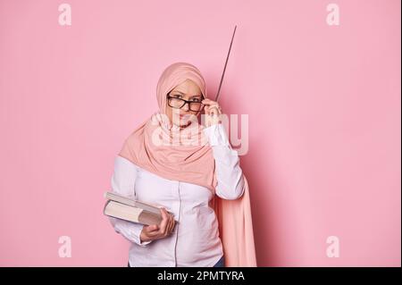 Donna incinta musulmana araba in hijab rosa, insegnante stressato guarda la macchina fotografica attraverso occhiali isolati su rosa Foto Stock