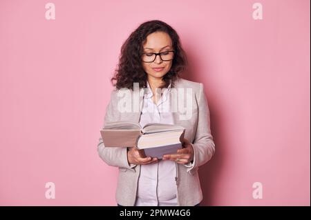 Donna incinta fiduciosa dai capelli scuri in abiti casual, indossando occhiali, leggendo libro di studio su sfondo rosa Foto Stock