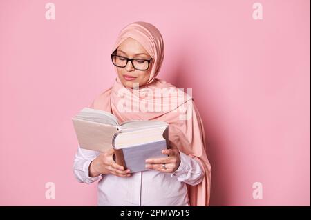 Bella donna musulmana del Medio Oriente in hijab rosa, libro di lettura isolato su sfondo a colori. Concetto di persone istruite Foto Stock