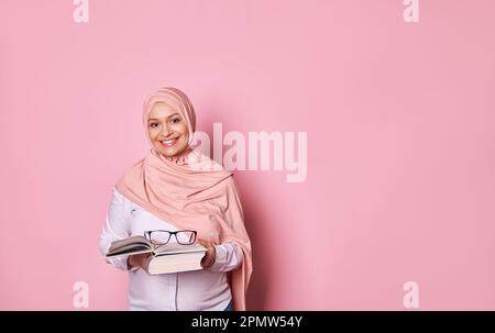 Donna incinta musulmana felice in hijab rosa, sorridente guardando la macchina fotografica, tenendo gli occhiali e libro. Islam ed educazione Foto Stock