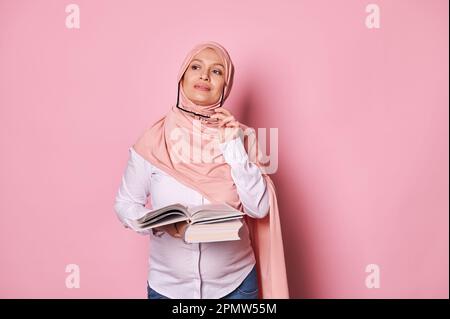 Donna incinta musulmana penosa in hijab, tenendo il libro sognatamente guardando via, ragionando sui suoi piani mentre congedo di maternità Foto Stock