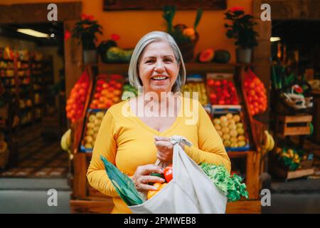 Vendita al dettaglio di alimenti a zero rifiuti - donna anziana felice che tiene un sacchetto riutilizzabile di verdure fresche presso il negozio del mercato locale - concetto ecologico sostenibile - Focus on face Foto Stock