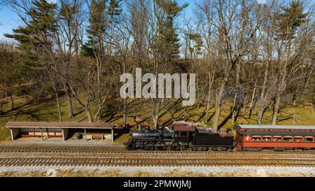 Una vista in drone di un treno a vapore restaurato che viaggia attraverso le Farmlands del RU che si sposta fino a una piccola stazione in un giorno d'autunno Foto Stock