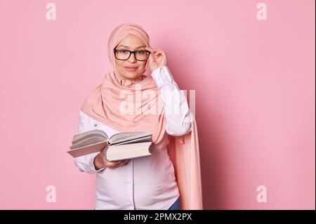 Fiducioso arabo musulmano educatore donna affascinante incinta in hijab rosa e occhiali, tenendo libro, guardando la fotocamera Foto Stock