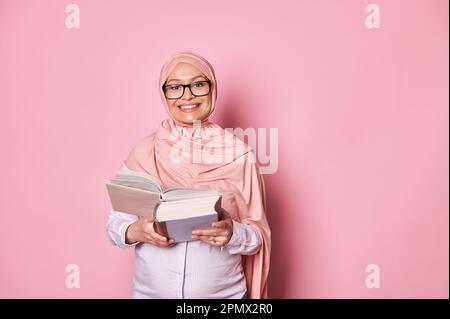 Sicura bella donna musulmana incinta in hijab rosa e gli occhiali alla moda, leggendo un libro, sorridendo guardando la macchina fotografica Foto Stock