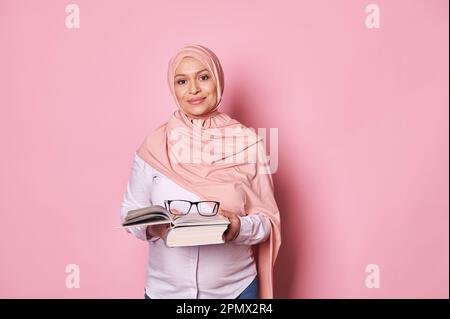 Donna musulmana fiduciosa educatore in hijab rosa, un insegnante di scuola che tiene occhiali e libro di studio, sfondo rosa Foto Stock