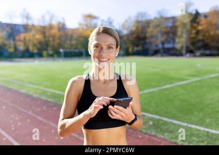 Giovane atleta runner in piedi in stadio con telefono e cuffie e brani per ascoltare musica, audiolibro, podcast. Sorridendo alla fotocamera. Foto Stock