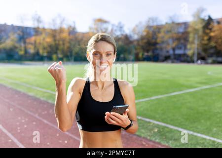 Giovane atleta che controlla il tempo al telefono dopo jogging, maratona. Mostra un gesto della mano di vittoria alla telecamera. Foto Stock