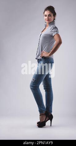 Un ritratto completo di una donna bianca che indossa tacchi alti, jeans blu e una blusa a maniche corte a righe. Si alza con la mano sull'anca, looki Foto Stock