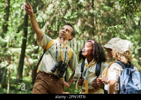 Gruppo vario di escursioni scout nella foresta con leader adulto che indica via illuminato dalla luce del sole Foto Stock