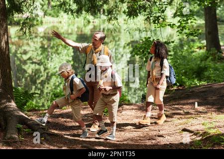 Vista laterale in un gruppo di scout diversi escursioni nella foresta con leader adulto che indica lontano dalla luce del sole Foto Stock