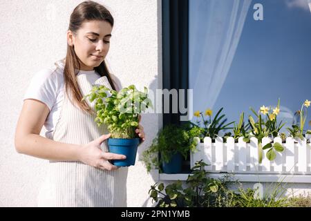 Bella donna in tute a strisce leggere reimpianto basilico in terrazza all'aperto. Giovane donna che si rilassa dopo una giornata di lavoro. Foto Stock