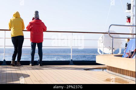 2 passeggeri della crociera si trovano sulla ringhiera a poppa di una nave da crociera e si affacciano sul mare aperto blu. Foto Stock