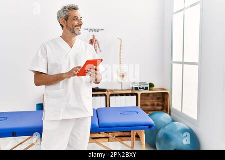 Fisioterapista uomo dai capelli grigi di mezza età sorridente sicuro usando il touchpad alla clinica di riabilitazione Foto Stock