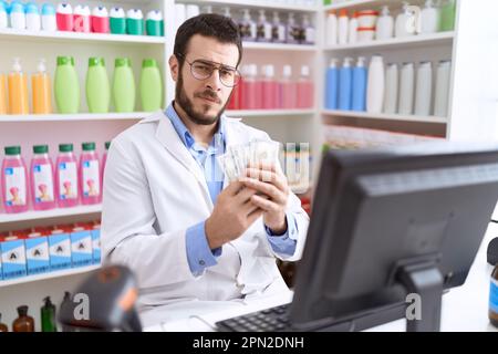 Farmacista giovane uomo ispanico che tiene dollari utilizzando il computer in farmacia Foto Stock
