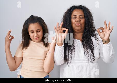 Madre e figlia in piedi su sfondo bianco rilassato e sorridente con gli occhi chiusi facendo gesto di meditazione con le dita. concetto di yoga. Foto Stock