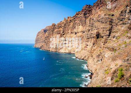 Le scogliere di Los Gigantes sovrastano la costa rocciosa della costa occidentale di Tenerife mentre una piccola barca a vela naviga attraverso le acque calme. Foto Stock
