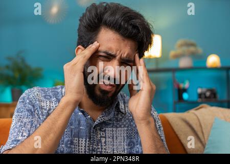 Deluso uomo indiano strofinare templi per curare problemi di mal di testa, che soffrono di tensione e emicrania, stress grimacing in dolore alta pressione sanguigna in casa appartamento camera. Indù ragazzo sul divano. Stili di vita Foto Stock