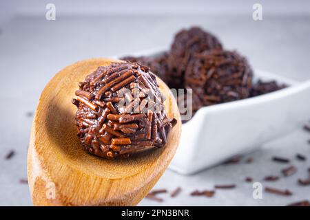 La caramella 'Brigadeiro' viene prodotta in un cucchiaio di legno con diversi dolci 'brigadeiro' su una ciotola bianca con spruzzate di cioccolato giacenti sul tavolo. Tipico Foto Stock