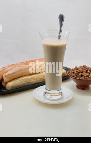 Un tavolo bianco con un bicchiere alto di horchata, un piatto pieno di fartoni e una ciotola con noci di tigre naturali su sfondo bianco e spazio copia Foto Stock