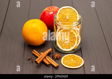 Scaglie di arancia in un vaso di vetro. Arancia cruda e mela fresca. Bastoncini di cannella e anice stellato sul tavolo. Spazio di copia. Sfondo di legno scuro. Foto Stock