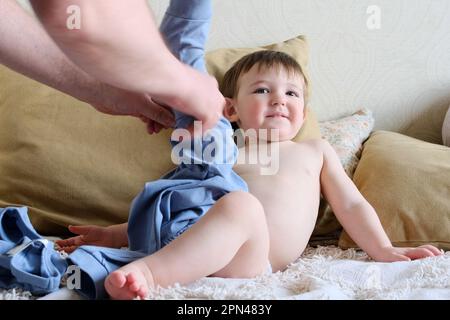 Il padre veste il bambino in un divano del pannolino. Papà uomo mette i vestiti sul bambino su un letto con cuscini. Bambino di circa due anni (un anno nove mesi) Foto Stock