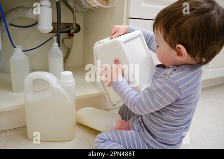 Un bambino gioca con prodotti chimici per la pulizia sotto il lavandino in cucina. Il bambino può contenere biberon con detergente. Bambino di circa due anni (uno yea Foto Stock
