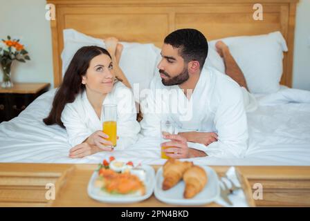 Coppia godendo la prima colazione sdraiata in Bed near Tray in Hotel Foto Stock