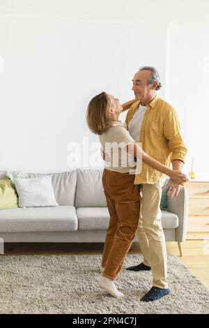 Sposi anziani amorevoli che si guardano con tenerezza mentre ballano insieme nel soggiorno di casa, tiro verticale Foto Stock