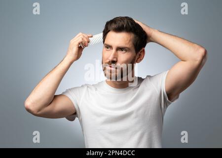 Concetto di Barbiere. Bell'uomo di mezza età in t-shirt bianca spazzolando i capelli, in piedi su sfondo grigio studio Foto Stock