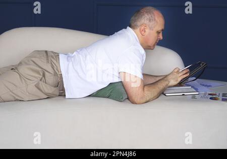 Un uomo d'affari si trova su un divano e guarda le tendenze dei dati su grafici e grafici in un tablet, prende appunti in un notebook. Lavoro a distanza Foto Stock