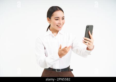 Ritratto della donna aziendale, chat video manager sul telefono cellulare, guarda lo smartphone e conduce una riunione online tramite l'app per smartphone, sfondo bianco Foto Stock