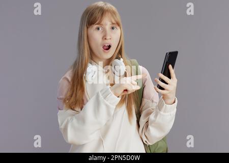 sorpresa moderna ragazza adolescente in tuta beige con zaino e cuffie utilizzando app per smartphone isolato su sfondo grigio. Foto Stock