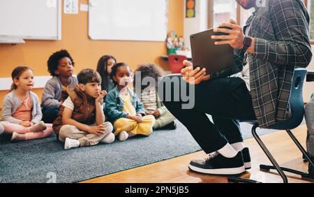 Insegnante di scuola elementare che usa un tablet per dare una lezione in classe. Educatore maschile è seduto davanti ai suoi studenti e mostra loro e vi educativo Foto Stock