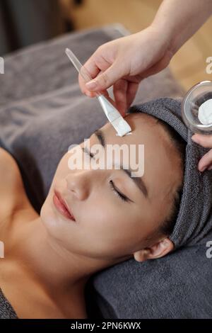 Cosmetologo applicare bianco maschera idratante del viso di giovane donna Foto Stock