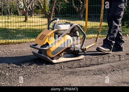 Vista ravvicinata del proprietario di casa utilizzare piastra compattatore strumento per costruire parcheggio con trucioli di asfalto riciclato in casa giardino all'aperto in primavera. Foto Stock