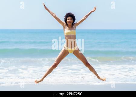 Donna etnica eccitata che salta con le braccia sollevate sulla spiaggia sabbiosa Foto Stock
