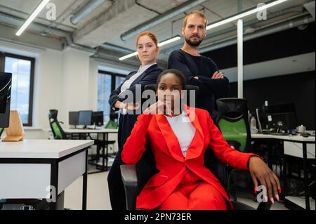 Donna caucasica dai capelli rossi, uomo caucasico bearded in piedi dietro una giovane donna afroamericana seduta in ufficio. Foto Stock
