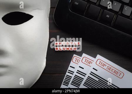 Concetto di whistleblowing: Maschera bianca su un tavolo di legno insieme a una tastiera per computer e documenti classificati Foto Stock