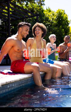 Felice gruppo diversificato di amici che hanno partito in piscina, tenendo tazze di plastica in giardino Foto Stock