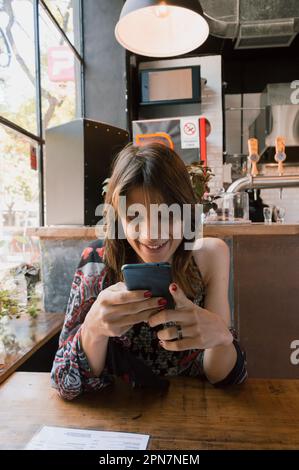 immagine verticale di giovane transgender donna latina di etnia argentina seduta in un ristorante, intrattenuto controllo di reti sociali al telefono, l Foto Stock