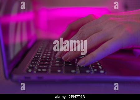 Persona che scrive al computer portatile di notte. La femmina usa la tastiera, scuote i pulsanti con le dita. Sicurezza su Internet, attacco informatico. Luce al neon alla moda. Crea Foto Stock