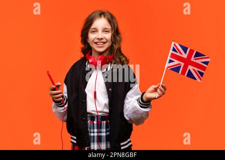 Teen in abiti casual e cuffie rosse con cellulare. La bambina della scuola tiene una piccola bandiera britannica. Studio all'estero concetto. Imparare l'inglese britannico con nativo Foto Stock