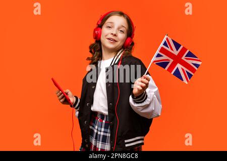 La bambina della scuola tiene una piccola bandiera britannica. Adolescenti in abiti casual e cuffie rosse con smartphone. Corsi di lingue online. Imparare l'inglese britannico. Ascolta Foto Stock